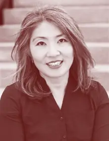 A portrait of May Young smiling, wearing a dark-colored blouse, and sitting on steps in an outdoor setting.