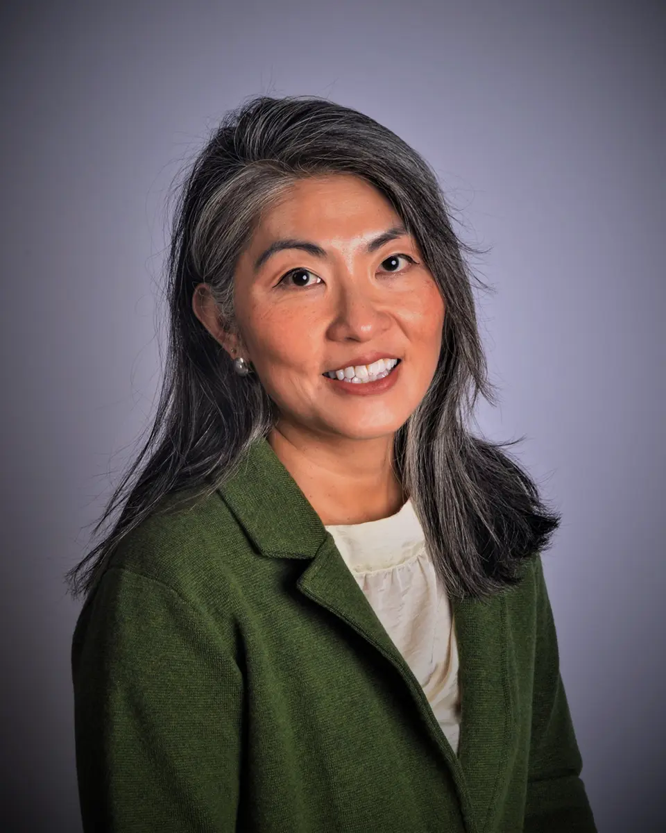 Portrait of May Young wearing a green blazer and smiling against a neutral gray background.